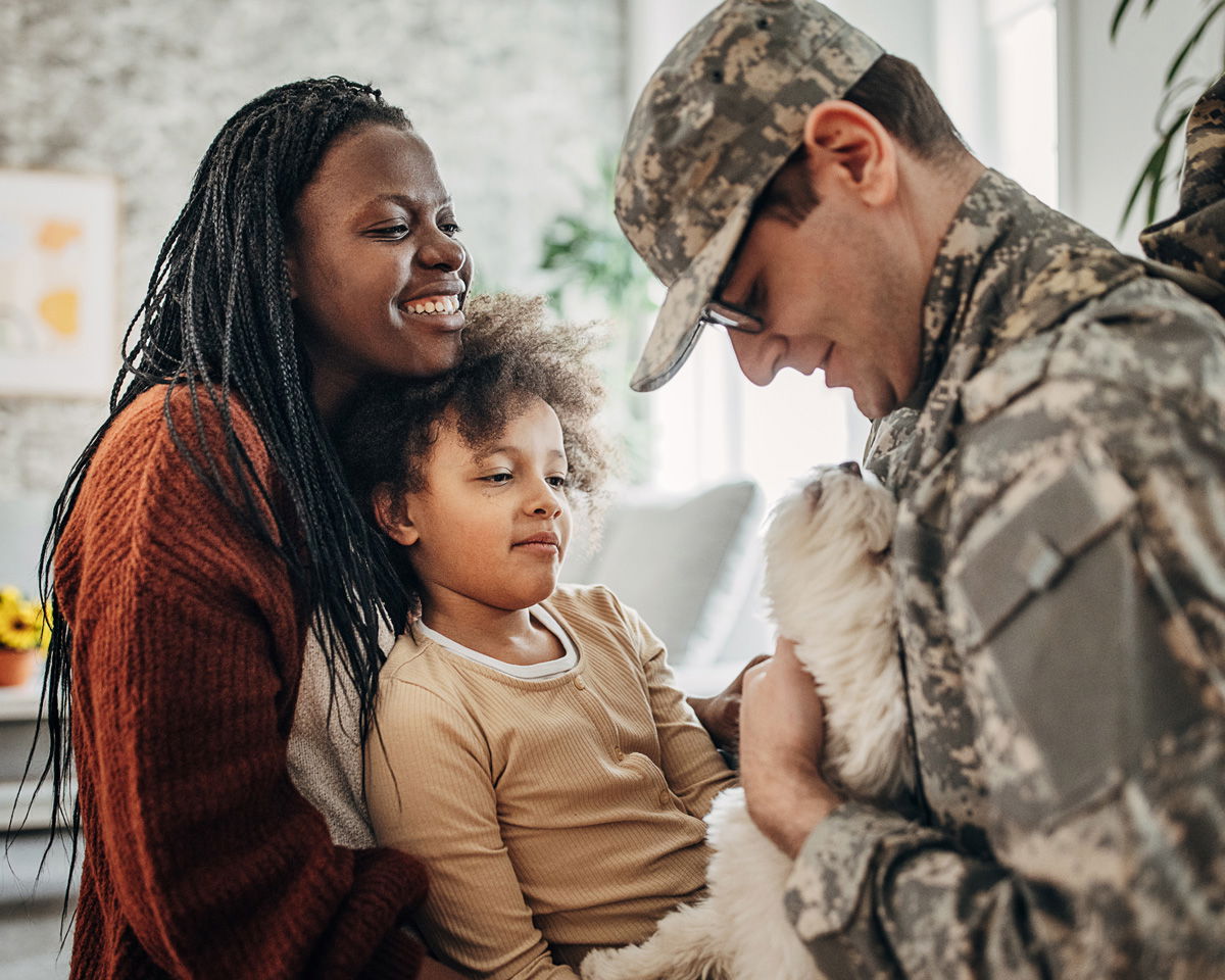 A smiling woman and child embrace a man in military uniform holding a small white dog. They appear happy in a cozy home setting. The man is wearing camouflage clothing and glasses.