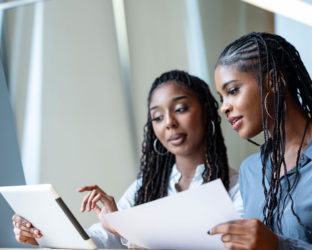 Two women with long braided hair are sitting and discussing something. One holds a sheet of paper, while the other points at a tablet. They appear focused and engaged in conversation. The background consists of light-colored walls.