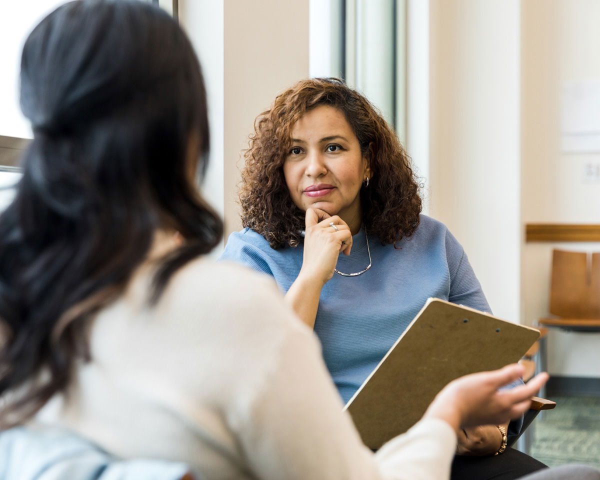A woman with curly hair, wearing a blue sweater, listens attentively with a clipboard in hand. She is seated across from another woman with long dark hair, who gestures as she speaks. They are in a sunlit room with large windows.