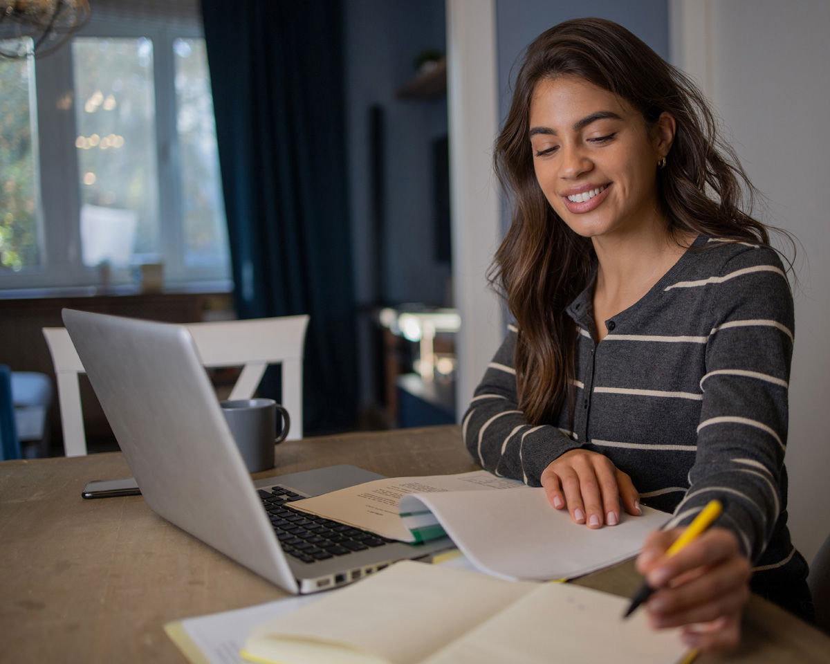 A smiling person with long hair is sitting at a desk, holding a yellow pen and writing in a notebook. A laptop and a mug are on the table. The background shows a window with natural light coming in.