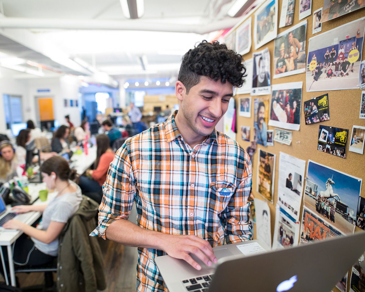 A person in a plaid shirt smiles while using a laptop in a busy office setting. In the background, people are working at desks, and a corkboard covered with photos is visible on the right.