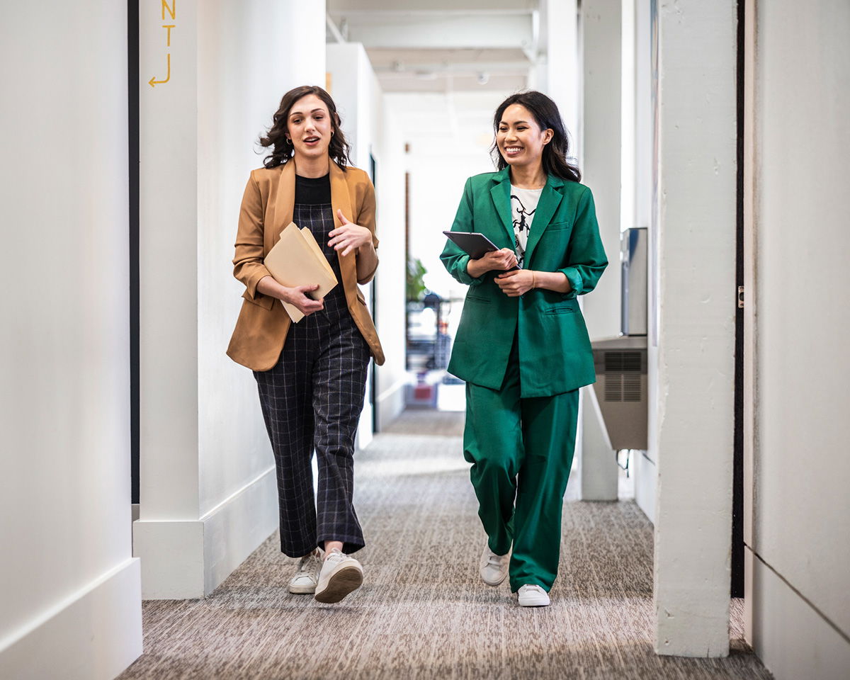 Two women walking in a hallway, wearing stylish suits. One is in a brown blazer, holding folders, and the other in a green suit, holding a tablet. They are smiling and appear engaged in conversation. The setting is a bright, modern office space.