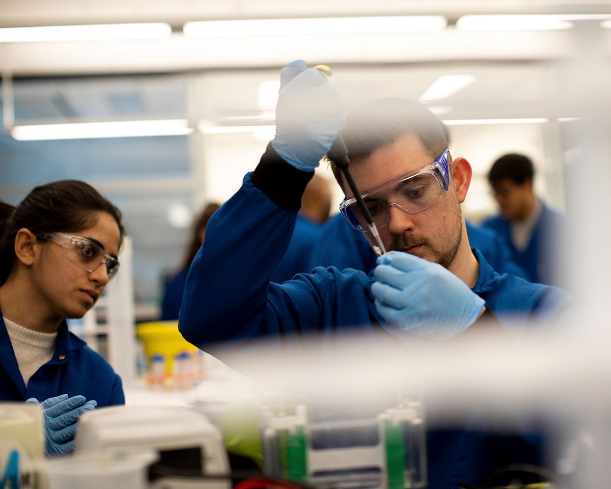 Two scientists in blue lab coats and safety goggles work in a laboratory. One is using a pipette to transfer liquid, while the other watches. The background is a well-lit lab space with equipment and other researchers.