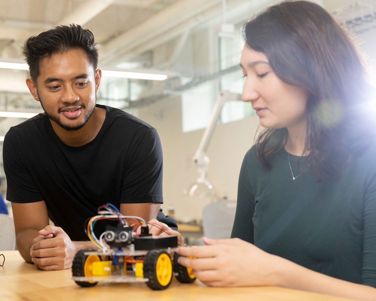 Two people in a workshop are focused on building a small wheeled robot on a table. The robot has wires and sensors visible. The background shows a well-lit, modern workspace.