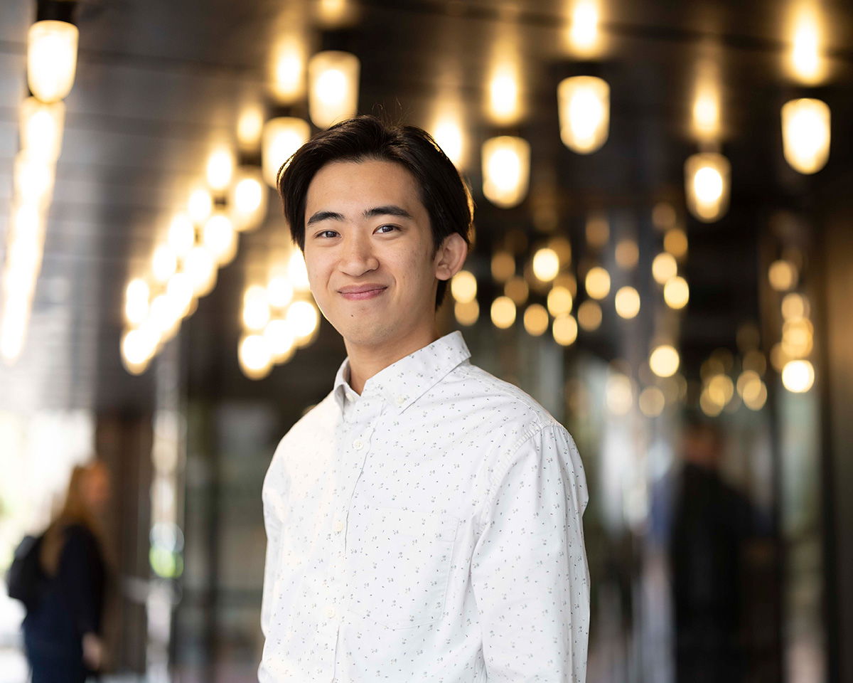 A person wearing a white shirt stands smiling in a well-lit corridor with numerous hanging lights. The background is softly blurred, highlighting the warm ambiance of the space.